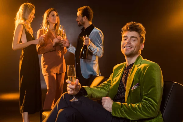 Man sitting on sofa and holding glass of champagne near multicultural friends on blurred black background — Stock Photo