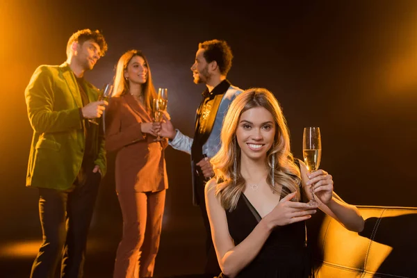 Mujer rubia feliz sentada en el sofá y sosteniendo una copa de champán cerca de amigos multiculturales sobre un fondo negro borroso - foto de stock