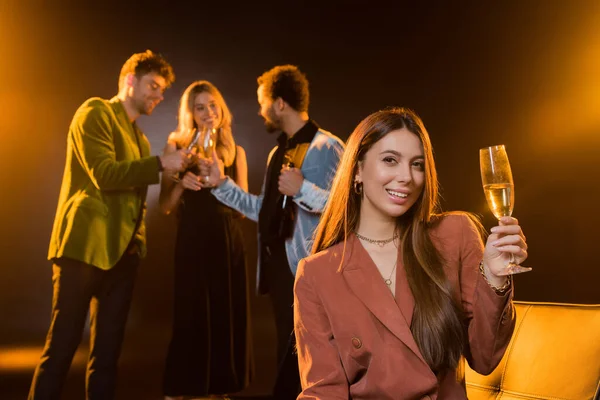 Feliz morena sentada en el sofá y sosteniendo una copa de champán cerca de amigos multiculturales sobre un fondo negro borroso - foto de stock