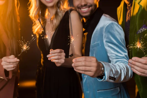 Cropped view of happy multicultural friends holding burning sparklers on black — Stock Photo