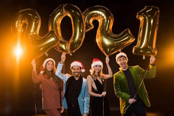 Happy multicultural friends in santa hats holding balloons with 2021 numbers on black — Stock Photo