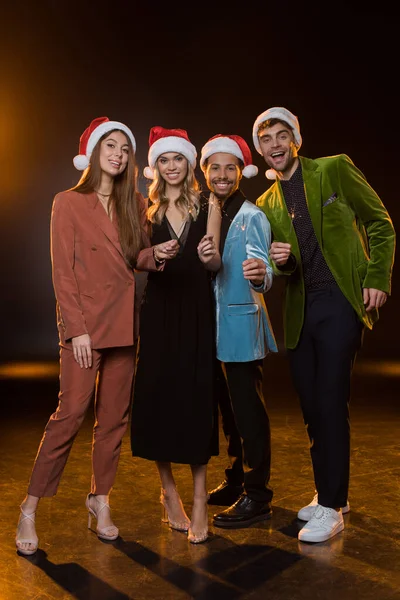 Pleine longueur de heureux amis multiculturels dans chapeaux Santa tenant des étincelles brûlantes sur noir — Photo de stock