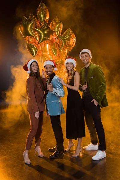 In voller Länge lächelnde multikulturelle Freunde in Weihnachtsmützen mit Gläsern mit Champagner in der Nähe von Luftballons auf schwarz mit Rauch — Stockfoto