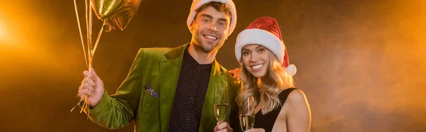 Coppia sorridente in cappelli di Babbo Natale che tiene bicchieri con champagne e palloncini su nero, banner — Foto stock