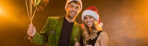 Sorrindo casal em chapéus santa segurando copos com champanhe e balões em preto, banner — Fotografia de Stock