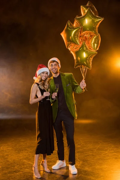 Full length of smiling couple in santa hats holding glasses with champagne and balloons on black — Stock Photo