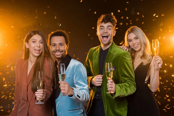 Smiling interracial friends holding glasses with champagne near falling confetti on black — Stock Photo