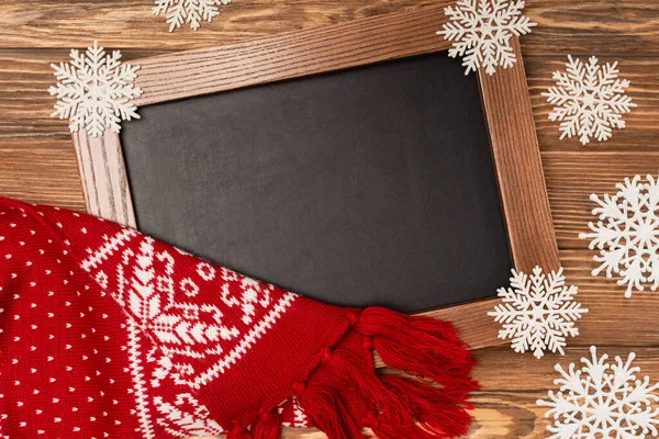 Top view of winter snowflakes on chalkboard and knitted scarf on wooden background — Stock Photo