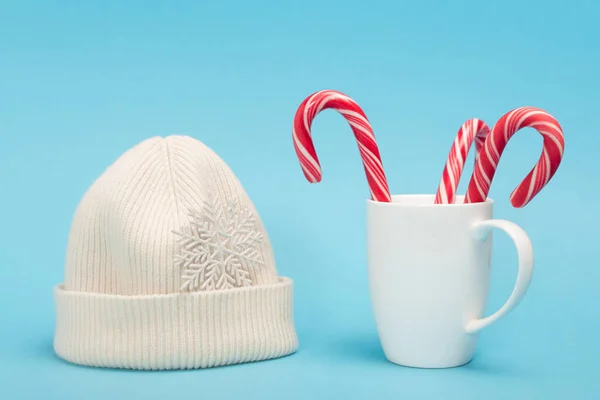 White beanie and candy canes in mug on blue background — Stock Photo