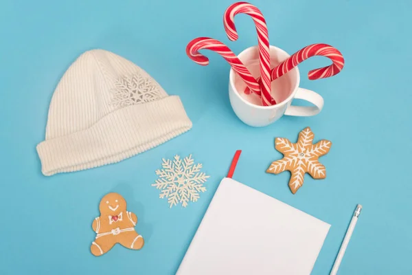 Top view of gingerbread cookies, beanie, candy canes and blank notebook on blue background — Stock Photo