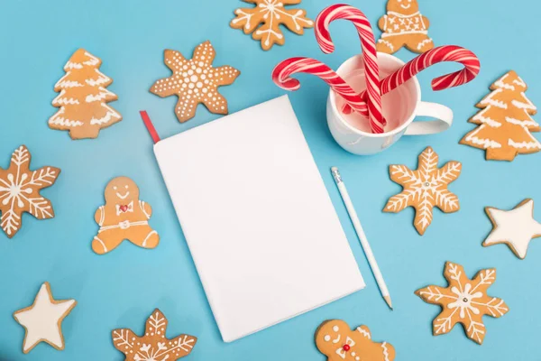 Ansicht von Lebkuchen, Zuckerstangen und leerem Notizbuch auf blauem Hintergrund — Stockfoto