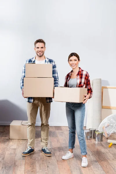 In voller Länge ein glückliches junges Paar, das in die Kamera schaut, während es zu Hause Kartons hält — Stockfoto