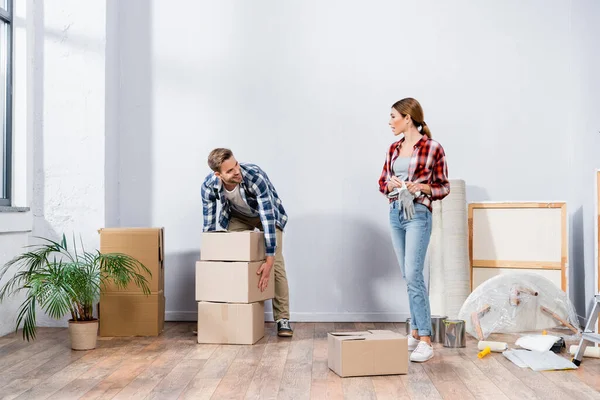 Homme souriant pleine longueur avec des boîtes en carton regardant femme tenant des gants à la maison — Photo de stock