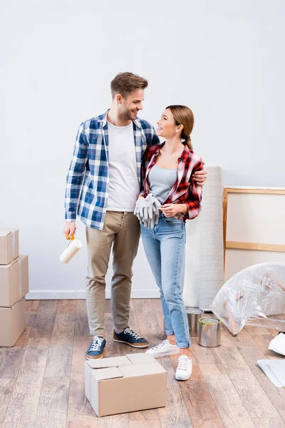 Larga duración de feliz pareja joven con guantes y rodillo de pintura mirándose mientras se abrazan en casa - foto de stock