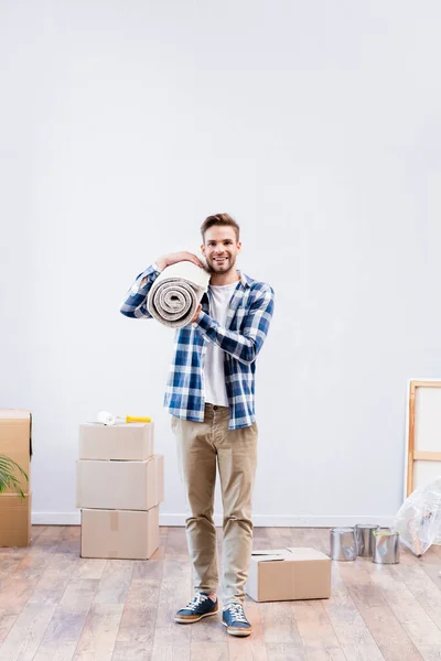 Full length of happy young man looking at camera while holding carpet roll near cardboard boxes and paint tins at home — Stock Photo