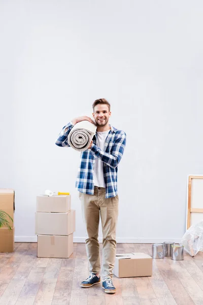 Volle Länge des glücklichen jungen Mannes, der in die Kamera schaut, während er Teppichrollen in der Nähe von Kartons und Farbdosen zu Hause hält — Stockfoto