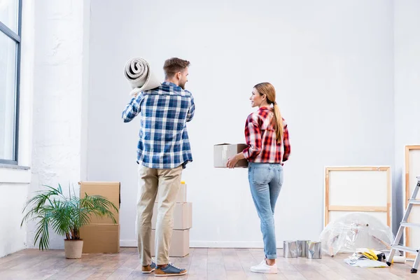 Back view of young couple holding cardboard box and carpet roll at home — Stock Photo