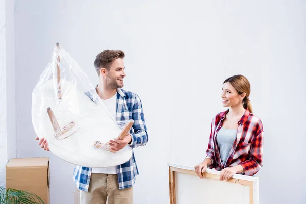 Smiling young man with coffee table covered with polyethylene looking at woman holding picture indoors — Stock Photo