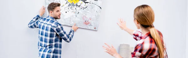 Young man looking at woman gesturing while removing picture from wall on blurred foreground at home, banner — Stock Photo