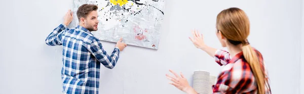 Joven hombre mirando a la mujer gesto mientras se quita la imagen de la pared en primer plano borrosa en casa, pancarta - foto de stock