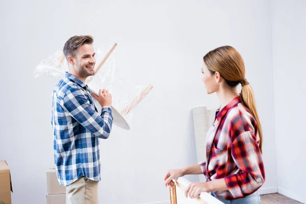 Smiling young man holding coffee table covered with polyethylene looking at woman with picture at home — Stock Photo