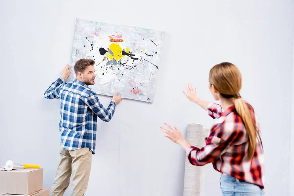 Young man looking at woman gesturing near picture at home on blurred foreground — Stock Photo