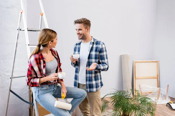 Lächelndes junges Paar mit Kaffeetassen im Gespräch nahe Leiter zu Hause — Stockfoto