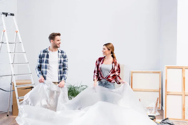 Heureux jeune couple avec du polyéthylène se regardant tout en couvrant la pièce pendant la rénovation de l'appartement — Photo de stock