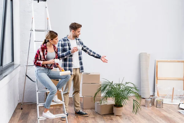 Junger Mann zeigt mit der Hand, während er Kaffeetasse in der Nähe von Frau hält, die während der Reparatur zu Hause auf Leiter sitzt — Stockfoto