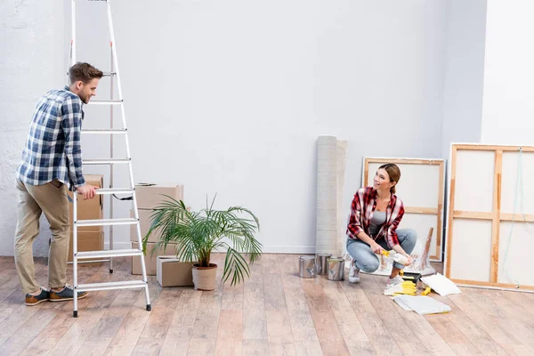 Lächelnder junger Mann blickt Frau mit Farbwalze an, während er sich zu Hause auf Leiter stützt — Stockfoto