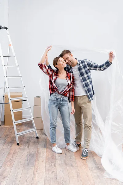 Full length of happy young couple hugging while covering with polyethylene near ladder at home — Stock Photo