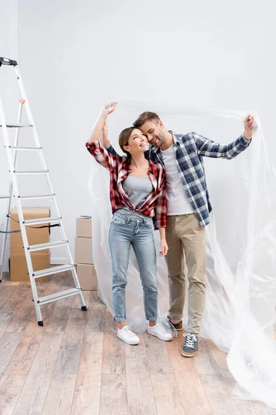 Pleine longueur de jeune couple heureux étreignant tout en couvrant de polyéthylène près de l'échelle à la maison — Photo de stock