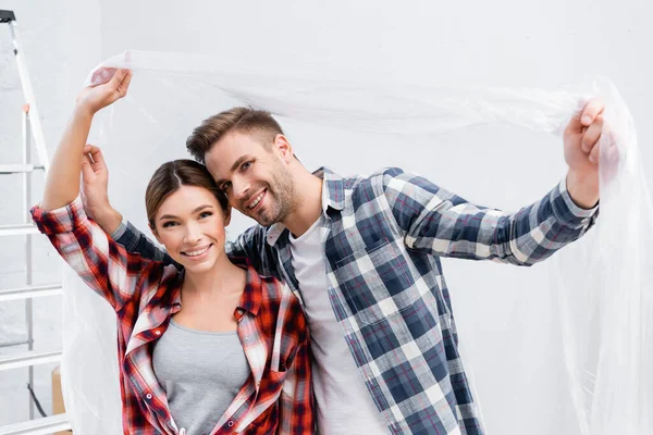 Feliz joven pareja mirando a la cámara mientras sostiene el polietileno en casa - foto de stock