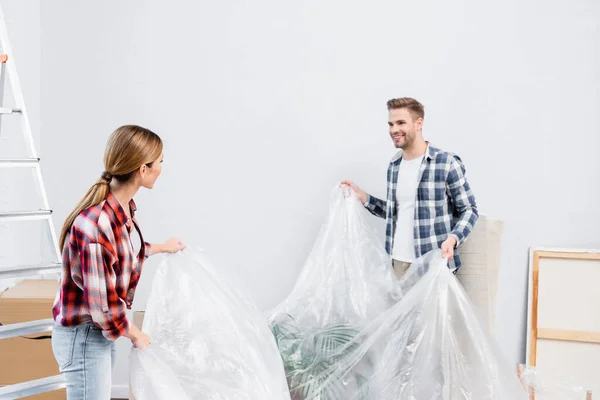 Feliz joven pareja cubriendo habitación con polietileno en casa - foto de stock