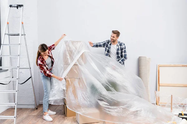 Full length of happy young couple with polyethylene covering cardboard boxes at home — Stock Photo