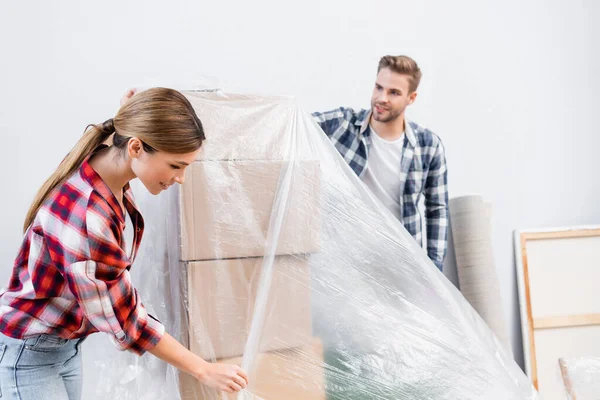 Smiling young couple with polyethylene covering cardboard boxes on blurred background at home — Stock Photo