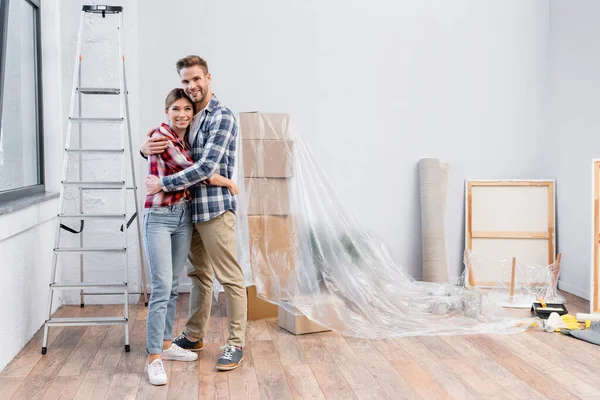 Volle Länge des glücklichen jungen Paares, das in die Kamera schaut, während es sich zu Hause umarmt — Stockfoto