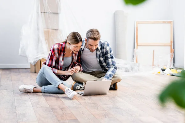Glückliches junges Paar blickt auf Laptop, während es zu Hause auf dem Boden auf verschwommenem Vordergrund sitzt — Stockfoto