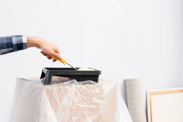 Cropped view of man putting paint roller in tray on box covered with polyethylene on blurred background indoors — Stock Photo