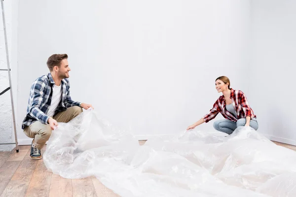 Happy young couple with polyethylene covering room while sitting on floor at home — Stock Photo