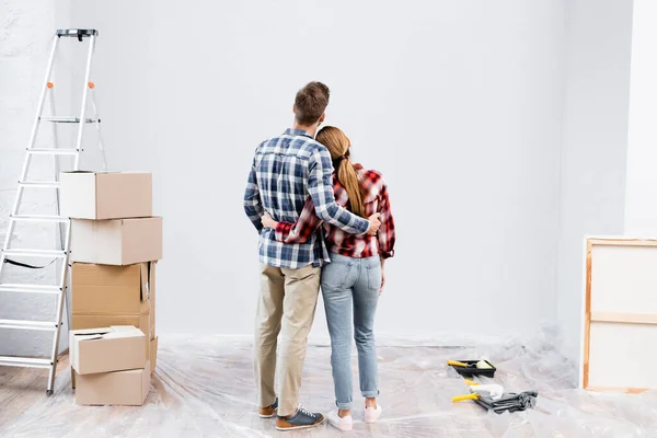 Back view of young couple hugging near ladder, cardboard boxes and paint rollers at home — Stock Photo