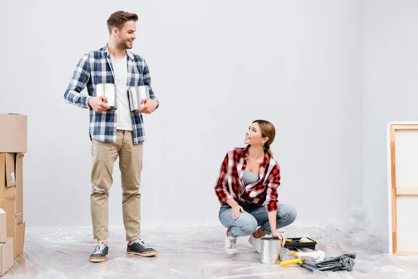 Pleine longueur de sourire jeune homme avec des boîtes de peinture regardant femme assise près des rouleaux de peinture sur le sol à la maison — Photo de stock