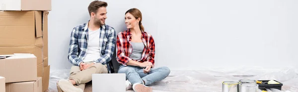 Sonriente joven pareja mirándose mientras se sienta cerca de la computadora portátil y cajas de cartón en el suelo en casa, pancarta - foto de stock