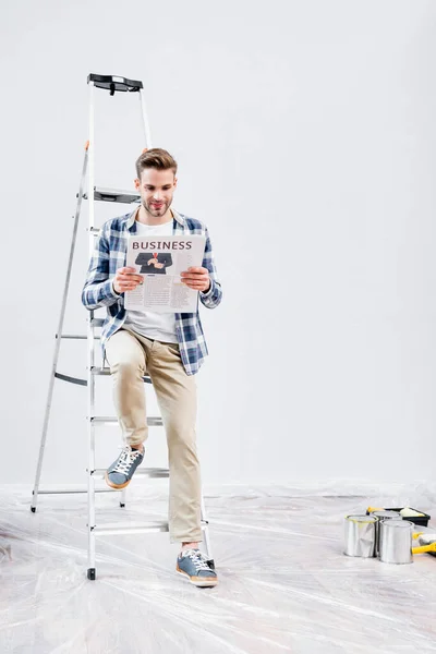 Toute la longueur du jeune homme souriant lisant le journal avec le lettrage d'affaires tandis que assis sur l'échelle près des boîtes de peinture à la maison — Photo de stock