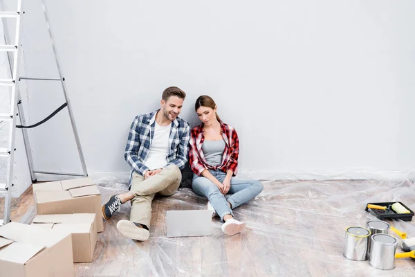 Full length of happy young couple looking at laptop while sitting on floor near cardboard boxes, tins of paint and ladder at home — Stock Photo