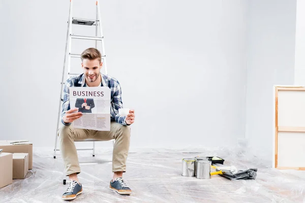 Voller Länge lächelnder junger Mann mit Kaffeetasse, der Zeitung liest, während er zu Hause auf Leiter sitzt — Stockfoto