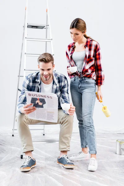 Full length of smiling young couple with coffee cups reading newspaper with business lettering near ladder at home — Stock Photo