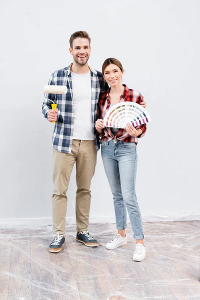 Pleine longueur de sourire jeune homme avec rouleau de peinture étreignant femme montrant palette de couleurs à la maison — Photo de stock