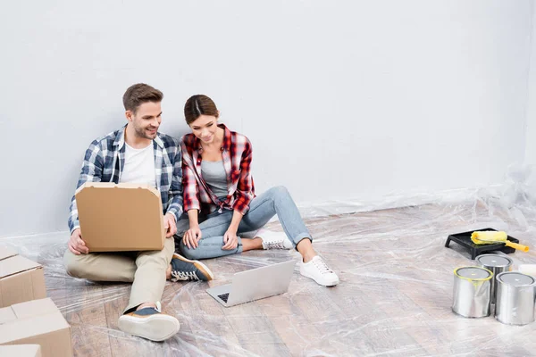 Volle Länge des lächelnden jungen Mannes mit Pappschachtel mit Pizza, der Film auf Laptop neben Frau anschaut, während er zu Hause auf dem Boden sitzt — Stockfoto