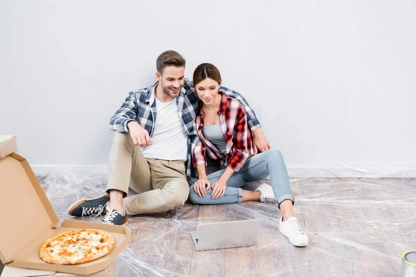 Longitud completa de feliz pareja joven abrazando y viendo la película en el ordenador portátil mientras está sentado en el suelo cerca de la caja con pizza en casa - foto de stock